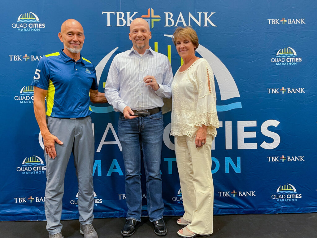 Ryan Carlson accepts the Carol J. Mindrup Award for outstanding volunteer service to the TBK Bank QC Marathon from founding race director Joe Moreno (left) and board member Tamia Keppy (right).