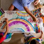 Family mother and kids drawing art picture on paper with multicolored chalks enjoy weekend leisure activity top view. Caring mom spending time with son and daughter happy motherhood relaxing at home childcare