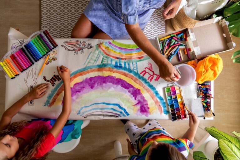 Family mother and kids drawing art picture on paper with multicolored chalks enjoy weekend leisure activity top view. Caring mom spending time with son and daughter happy motherhood relaxing at home childcare